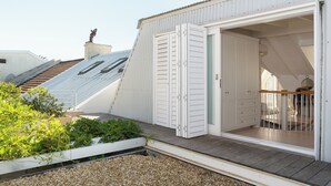A white building with a sloped roof, a wooden deck, and a sliding glass door leading to an interior with a staircase and a small balcony.