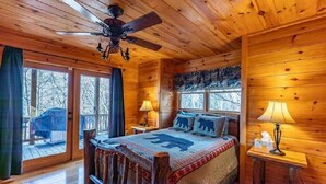 A cozy cabin room with a wooden ceiling, a bed with a bear-themed blanket, two bedside lamps, and a view of the outside through large windows.