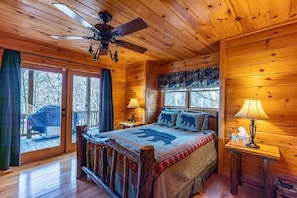 A cozy cabin room with a wooden ceiling, a bed with a bear-themed blanket, two bedside lamps, and a view of the outside through large windows.