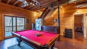 A wooden interior with a pool table, a staircase, and a fireplace.