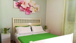 Een slaapkamer met een wit bed, groen beddengoed en een nachtkastje met een lamp.
