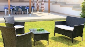 Een buitenterras met rieten meubels en een tafel, een overdekt zitgedeelte met tafels en stoelen, en een trap naar een hoger gelegen niveau.