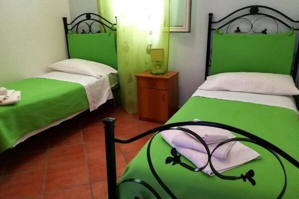 A room with two beds, each with green bedding and black headboards. There is a wooden nightstand between the beds with a lamp on it.