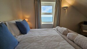 Ein ordentlich bezogenes Bett mit weißen Leintüchern und zusammengerollten Handtüchern, eine Nachttischlampe und Blick auf das Meer durch ein Fenster mit Vorhängen.