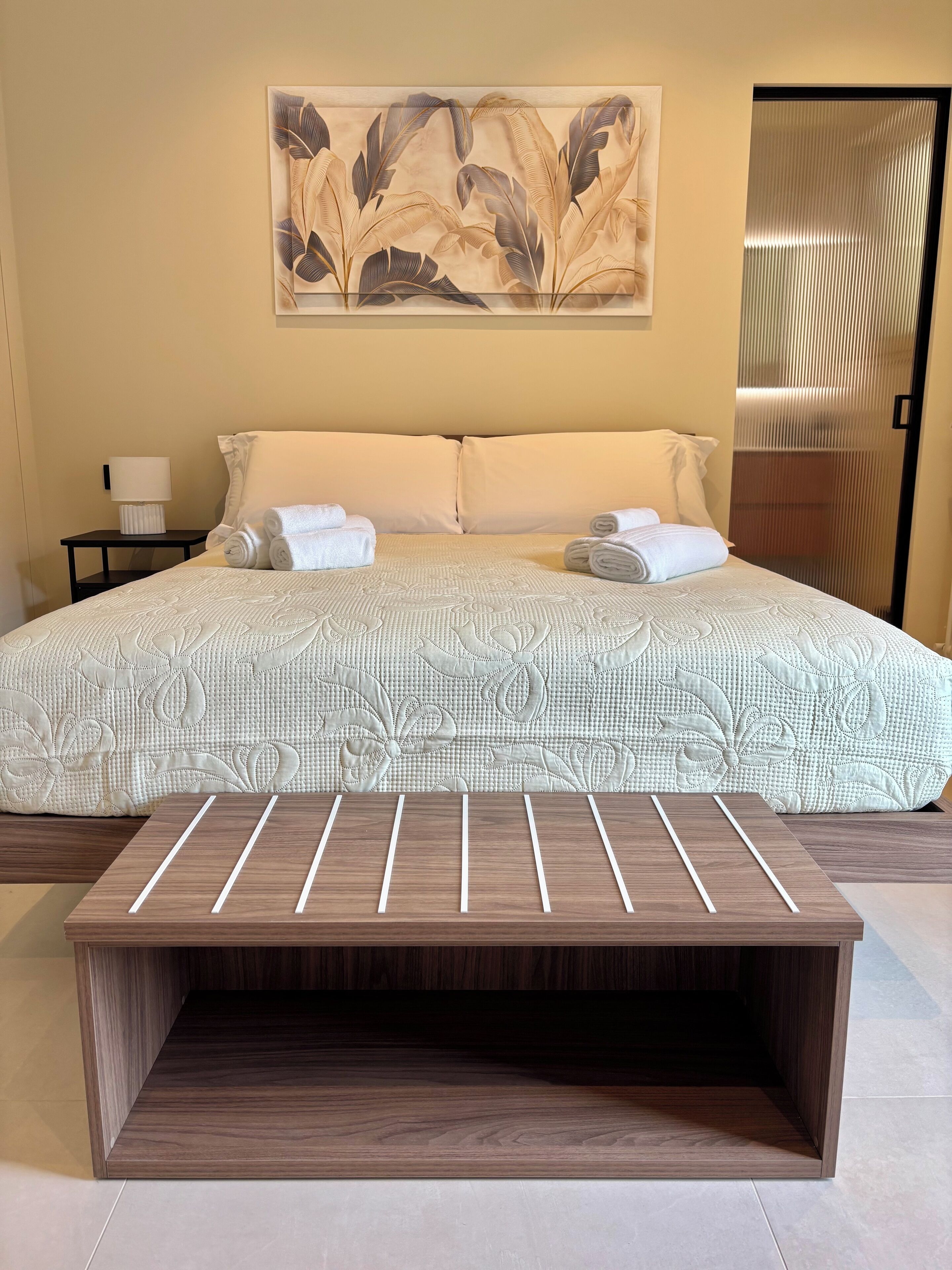 A neatly made bed with a floral-patterned bedspread, two pillows, and rolled towels on a nightstand.