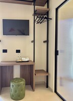 A modern room with a wooden desk, a green ottoman, a TV mounted on the wall, and a vertical rack with hangers.