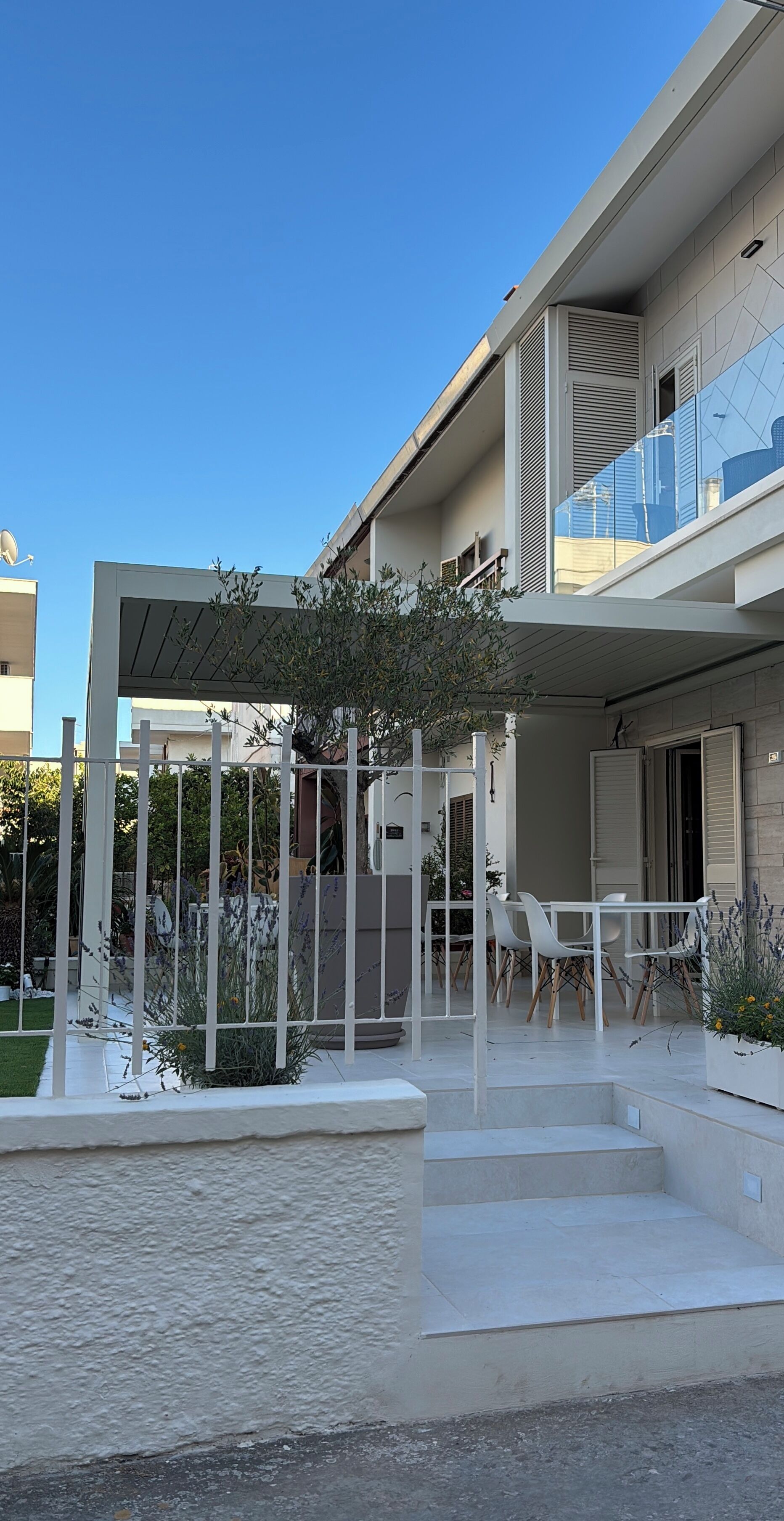 Un immeuble résidentiel moderne doté d’un espace extérieur aménagé et abrité, d’un escalier menant à l’entrée et d’un jardin avec des plantes et une clôture blanche.