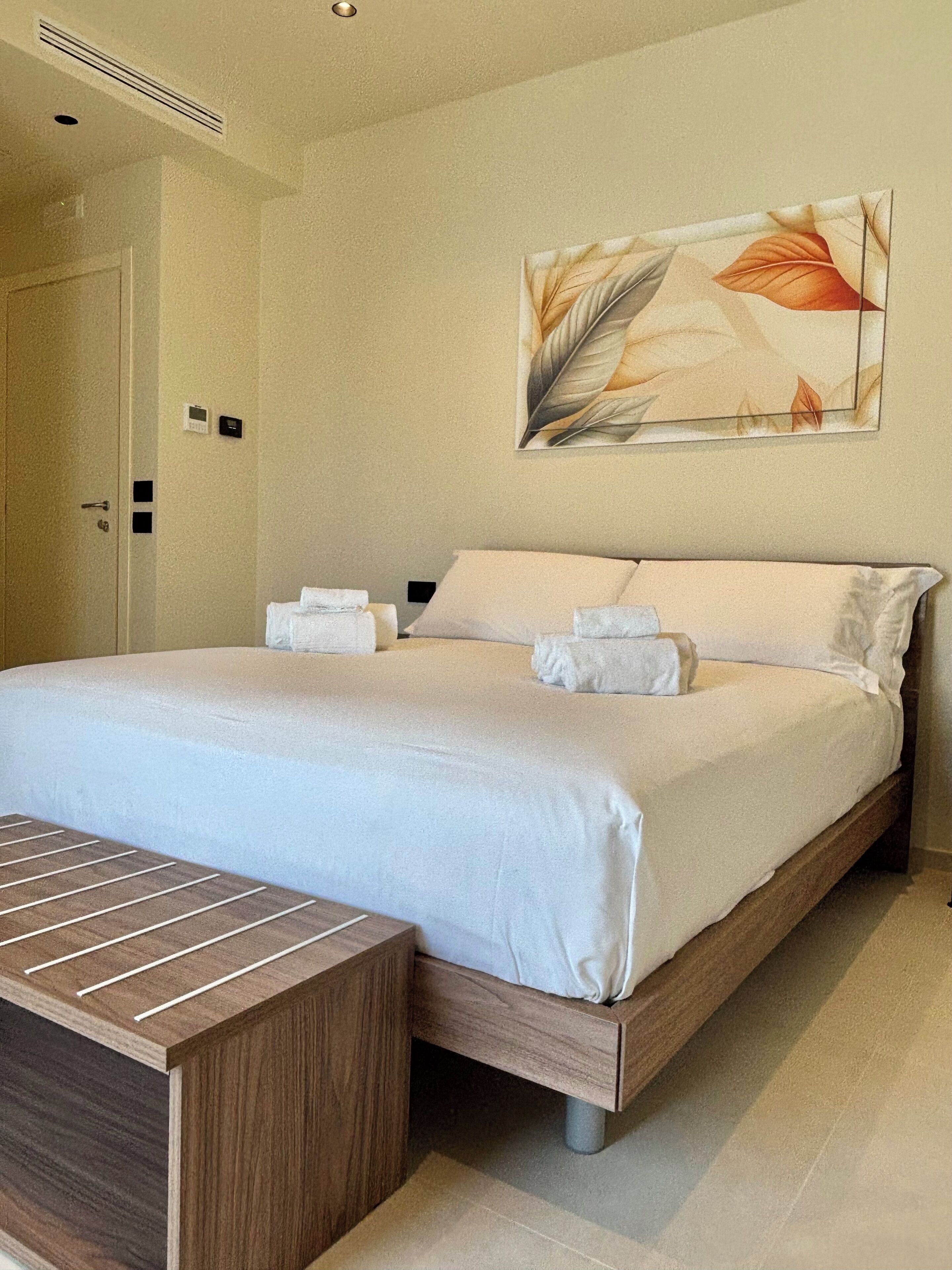 A neatly made bed with white linens and pillows, a wooden bench, and a wall-mounted artwork of leaves.