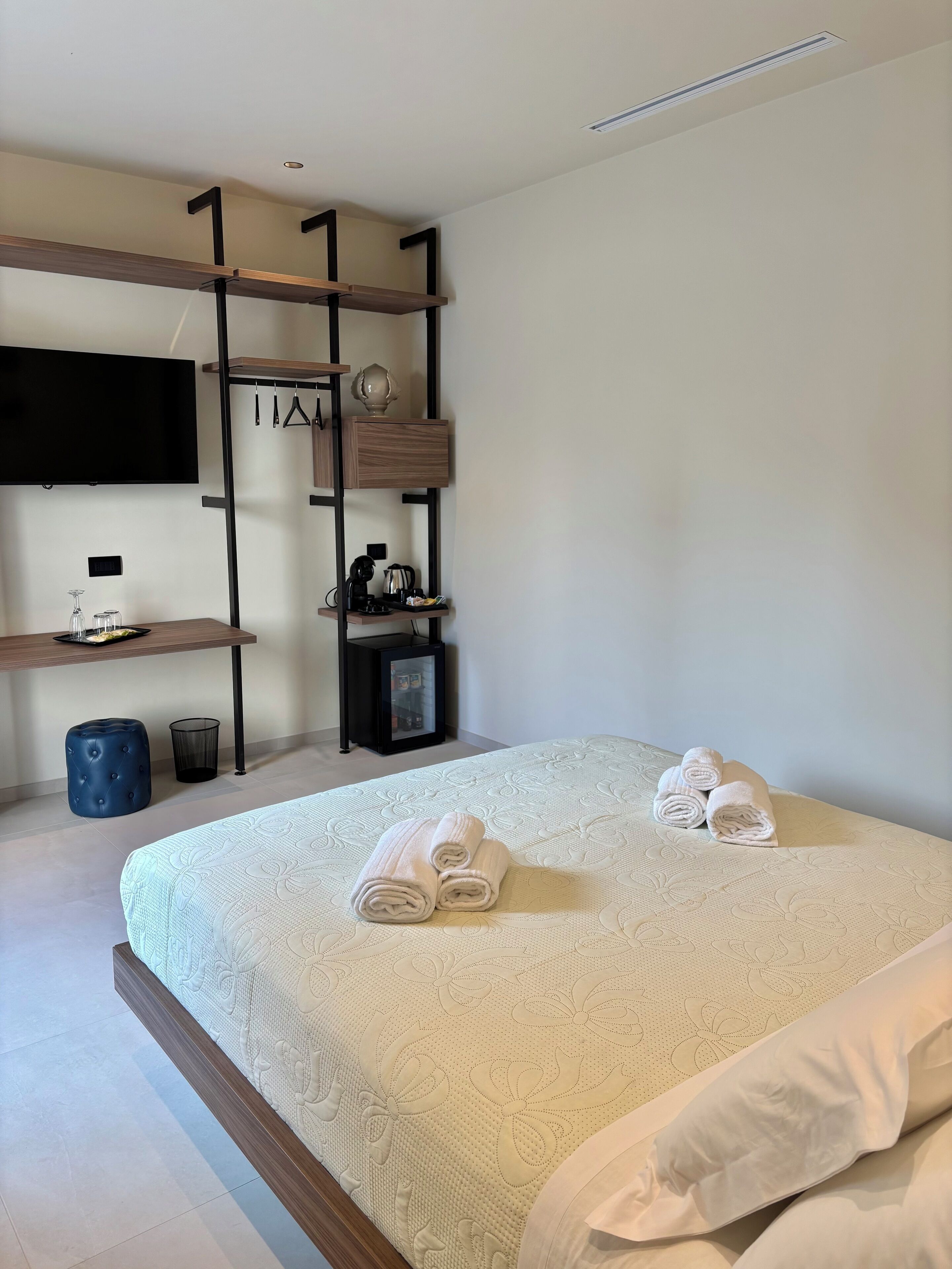 Un dormitorio ordenado con cama, televisor montado en la pared, una mesita con hervidor y estante con un objeto decorativo.
