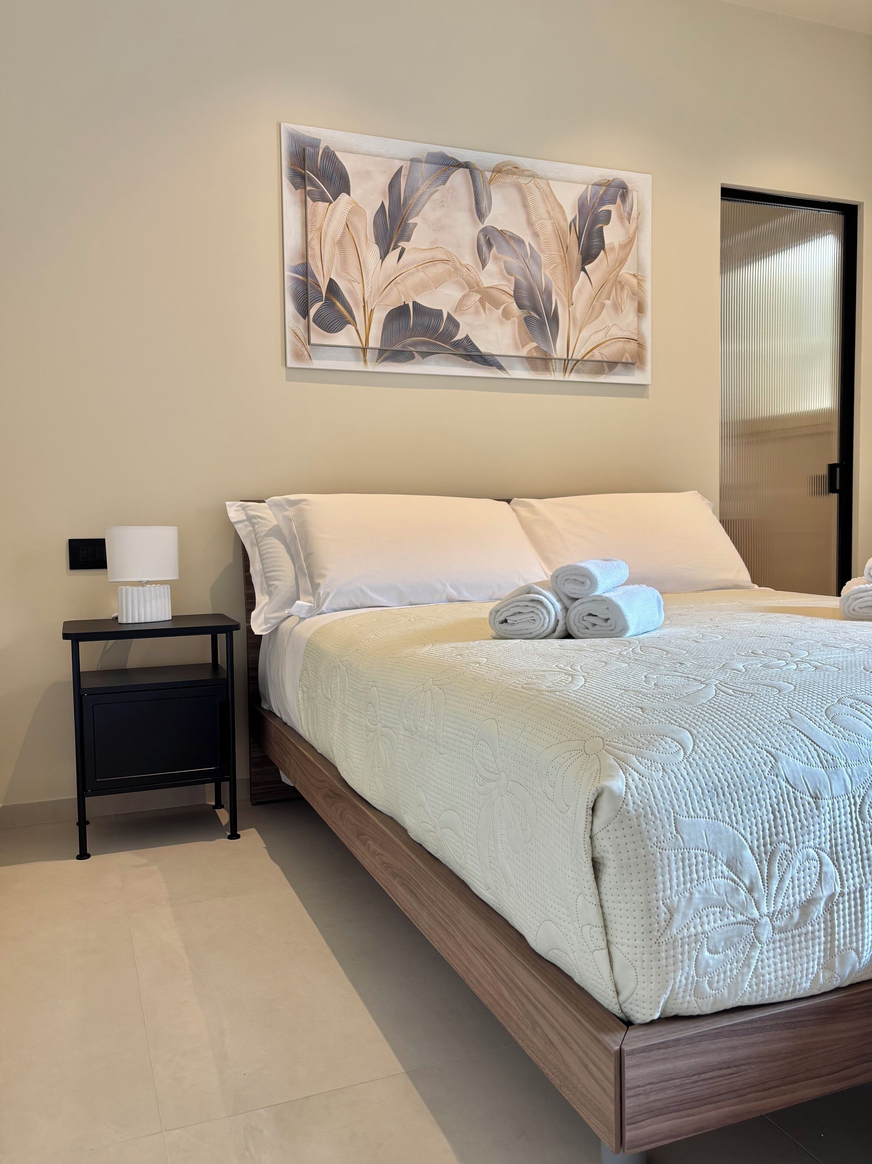 Un letto rifatto con un piumone bianco e asciugamani arrotolati, un comodino con una lampada e un'opera d'arte a parete raffigurante foglie.