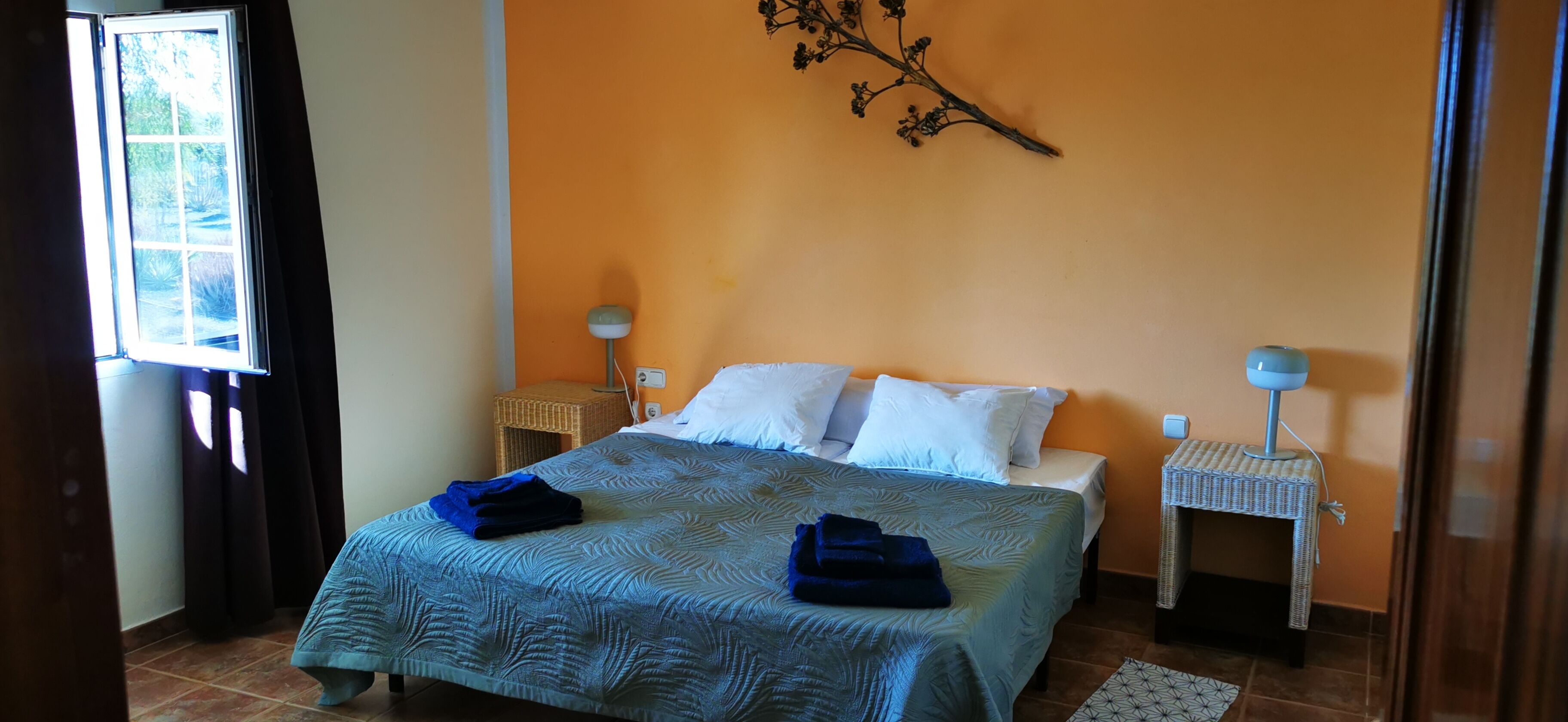 Un llit ben arrenjat amb un estovat estampat i dos coixins, una tauleta de nit amb una làmpada, i una decoració de branca penjada a la paret.