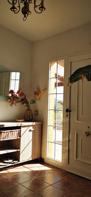 A room with a wooden cabinet, a vase with flowers, a mirror, and a door with a view of the outside.