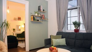 Una sala de estar con un sofá gris oscuro, una mesa redonda con fruta, una estantería y una ventana con cortinas.