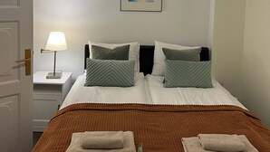 Cama bien hecha con dos almohadas, una manta marrón y dos toallas dobladas. Hay una lámpara de noche en un mesita, un cuadro en la pared y una lámpara colgante sobre la cama.
