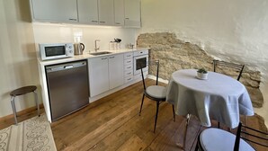 Eine kleine Küche mit weißen Schränken, einer Mikrowelle, einem Wasserkocher und einem Waschbecken. Ein runder Tisch mit weißem Tischläufer und zwei Stühle stehen bereit. Der Boden ist aus Holz, und an einer Wand befindet sich Naturstein.