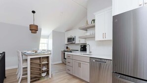 A modern kitchen with white cabinetry, a stainless steel refrigerator, and a dining area with a white table and chairs. - Downtown Newport Penthouse (Newport)