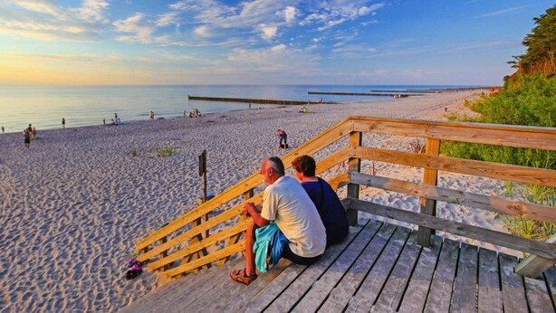 Eine hölzerne Mole mit zwei Personen, die auf ihr sitzen und auf einen sandigen Strand mit ruhiger See und blauem Himmel blicken.