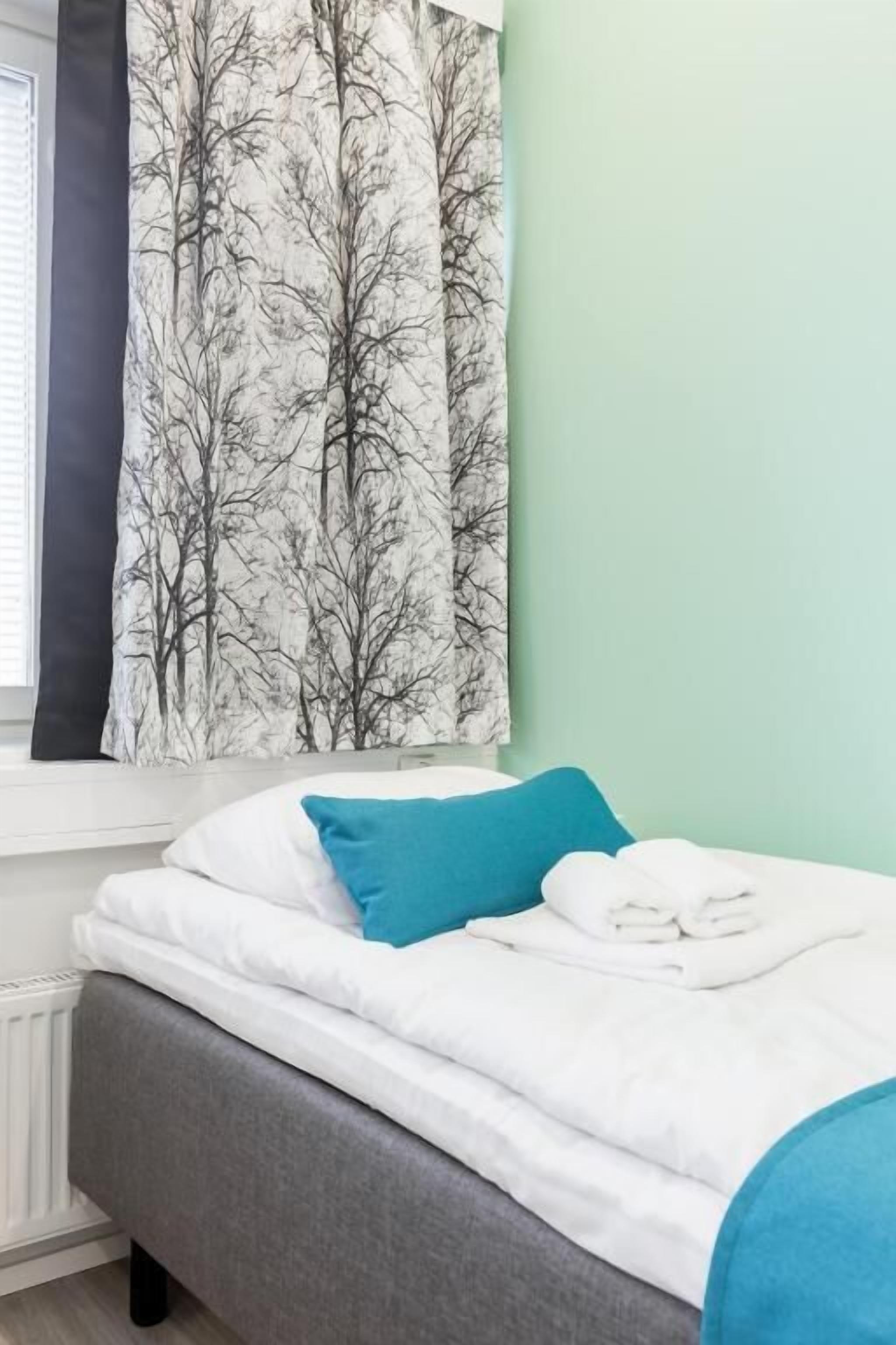 Una cama bien tendida con ropa de cama blanca y azul, un banco tapizado en gris y una ventana con cortina negra con estampado de árbol.