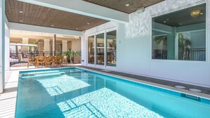 Une piscine avec une terrasse en bois, des portes vitrées donnant sur un espace extérieur aménagé pour se détendre, et une vue sur les environs.