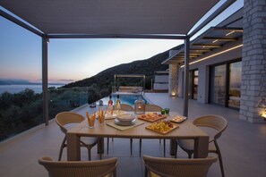 A dining area with a view of a pool and hills, featuring a table set with plates, glasses, and cutlery. - Efxenia Villas-Villa Luna (Lefkada)
