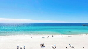 A beach with white sand, a clear blue sky, and a pier in the distance.