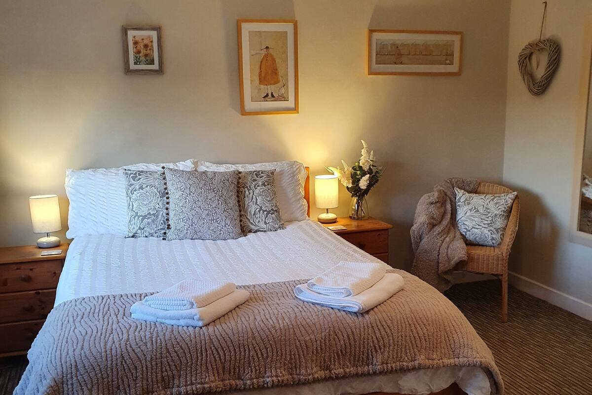 A neatly made bed with a patterned bedspread and pillows, a bedside table with a lamp, and a chair with a cushion.