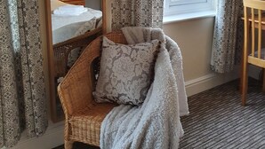 Een hotelkamer met een grote spiegel, een rieten stoel met een plaid, een houten ledikantje, kamer met behang en een raam met uitzicht.