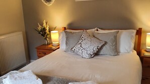 Een net opgemaakt bed met een zachte grijze deken en twee gevouwen witte handdoeken, een ingelijst kustlandschap erboven, een decoratief hart aan de muur, en een klein tafeltje met een lamp en een bloevas naast het bed.