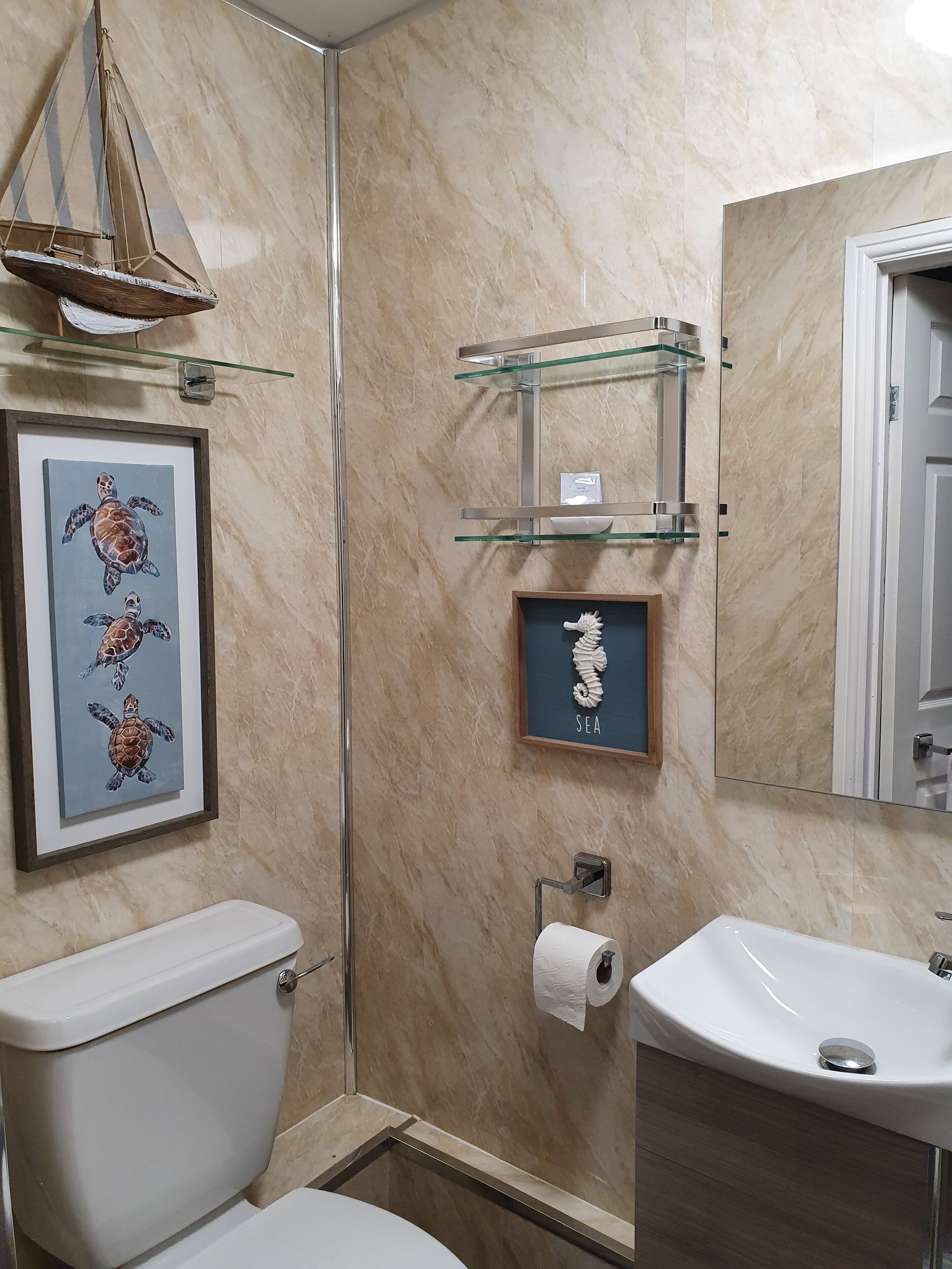 A bathroom with a marble wall, a framed sea turtle artwork, a toilet, a sink, and a glass shelf with toiletries.