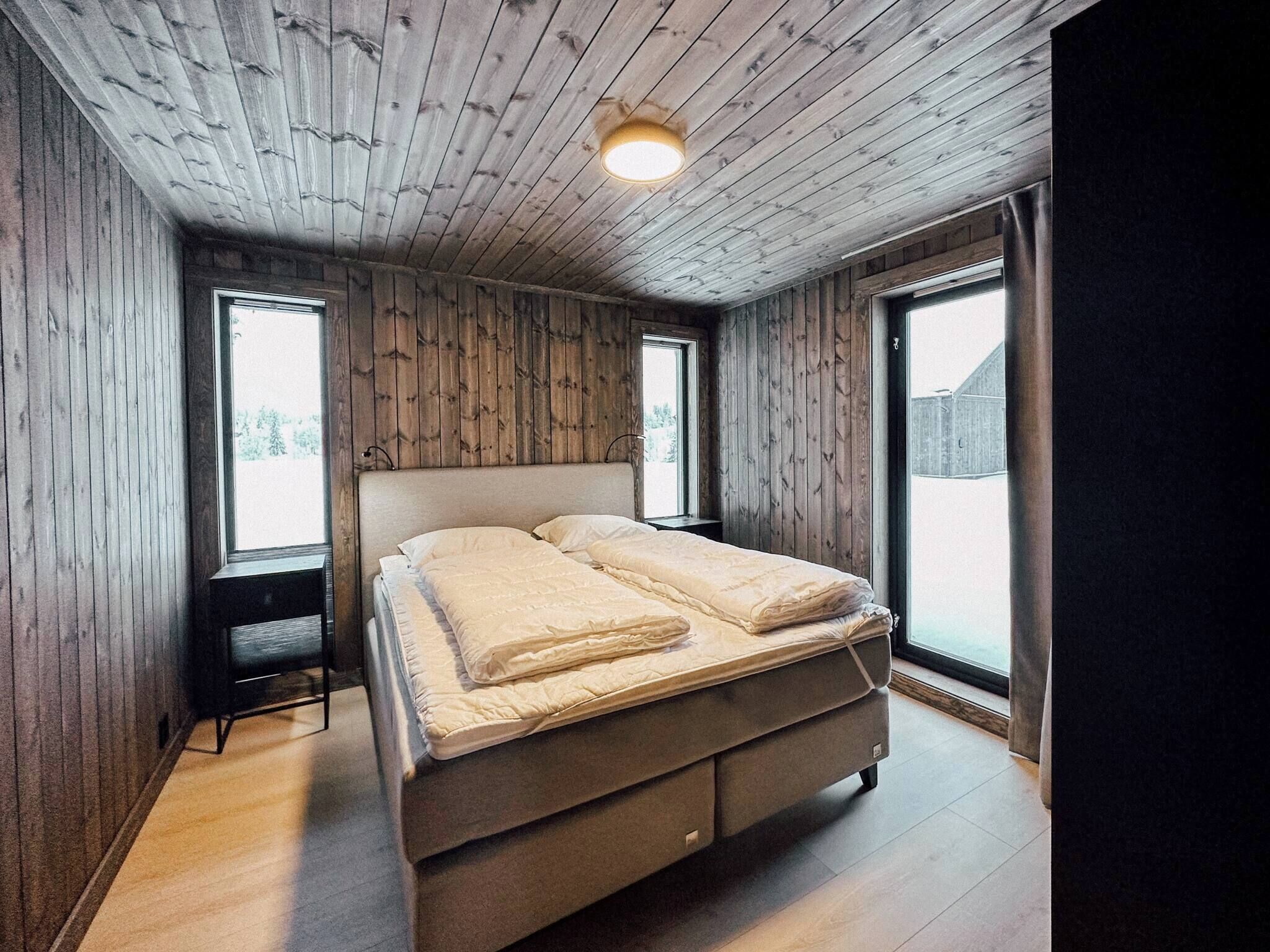 A bedroom with a wooden ceiling, a bed with beige bedding, two bedside tables, and large windows.