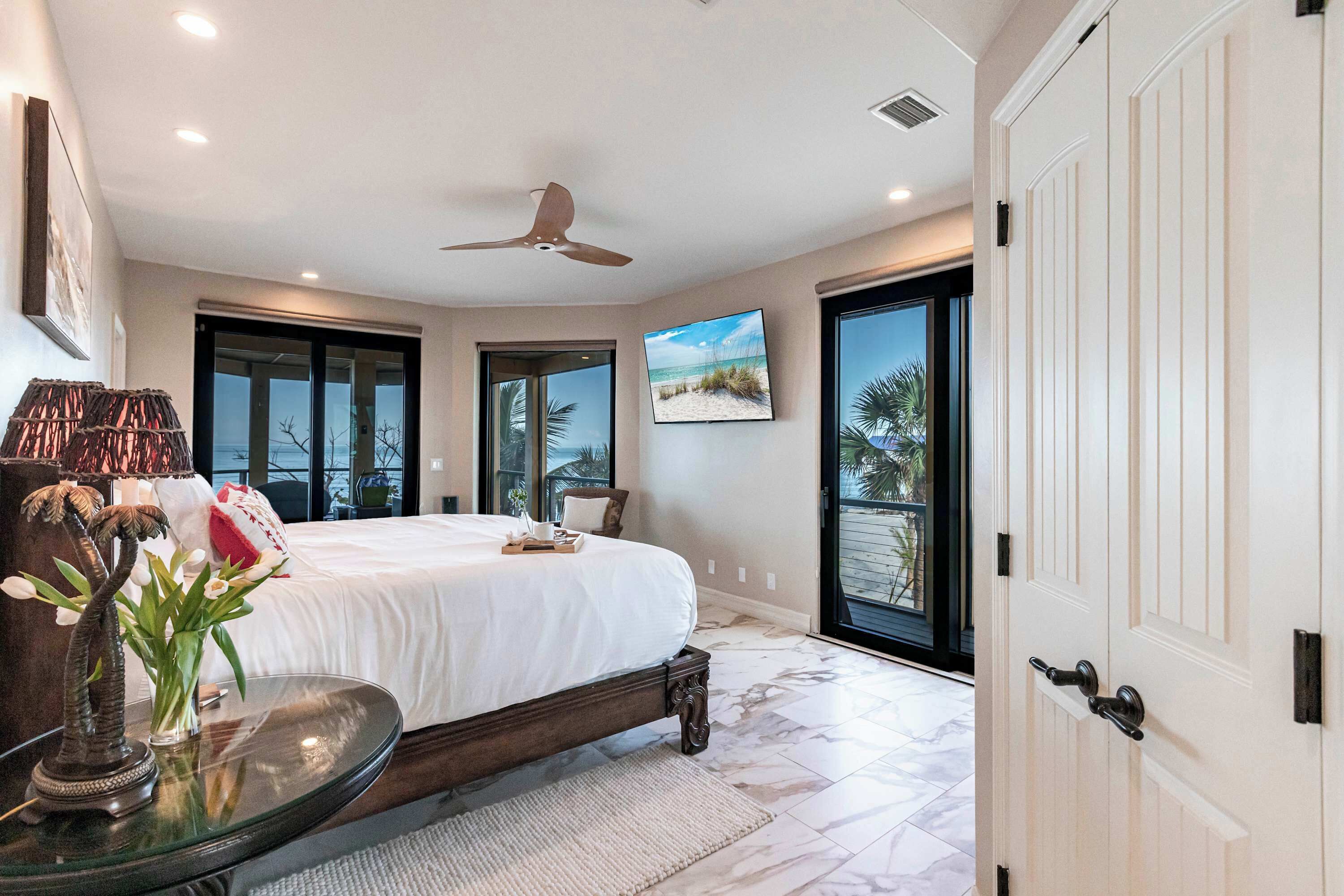 A bedroom with a large bed, a ceiling fan, a TV, and a view of the outdoors.