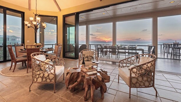 Amplo espaço interno com vista para o pôr do sol sobre a água, com móveis de vime, mesa de centro de madeira e mesa de jantar com cadeiras.