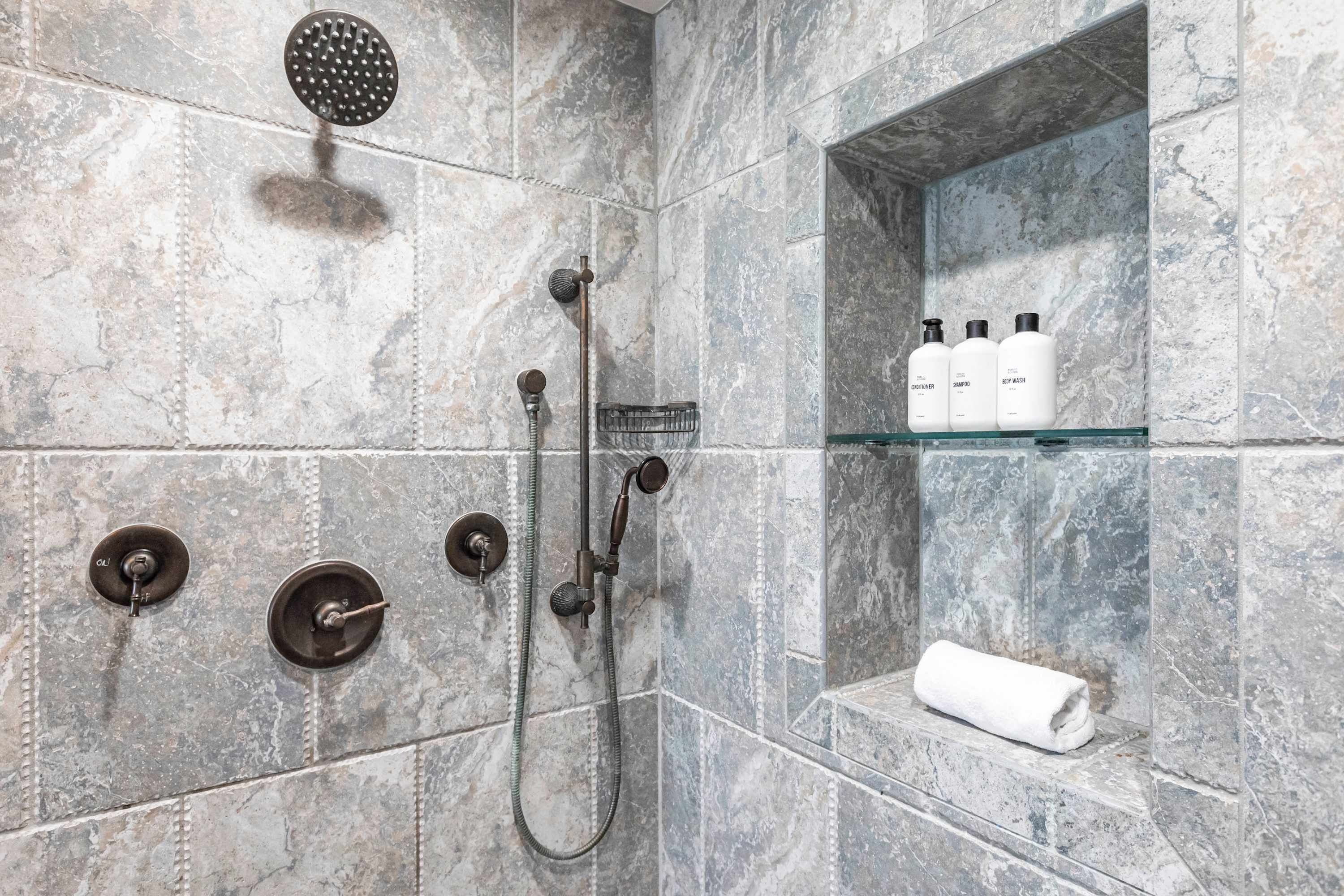 Eine moderne Dusche mit Handbrause, Deckenbrause und Regendusche. Eine Glasablage mit Pflegeprodukten und einem gerollten Handtuch.