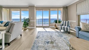 Ein geräumiges Wohnzimmer mit einer großen Schiebeglastür, die auf einen Balkon mit Meerblick führt. Zwei Sofas, ein Couchtisch und ein Blumentopf sind vorhanden.