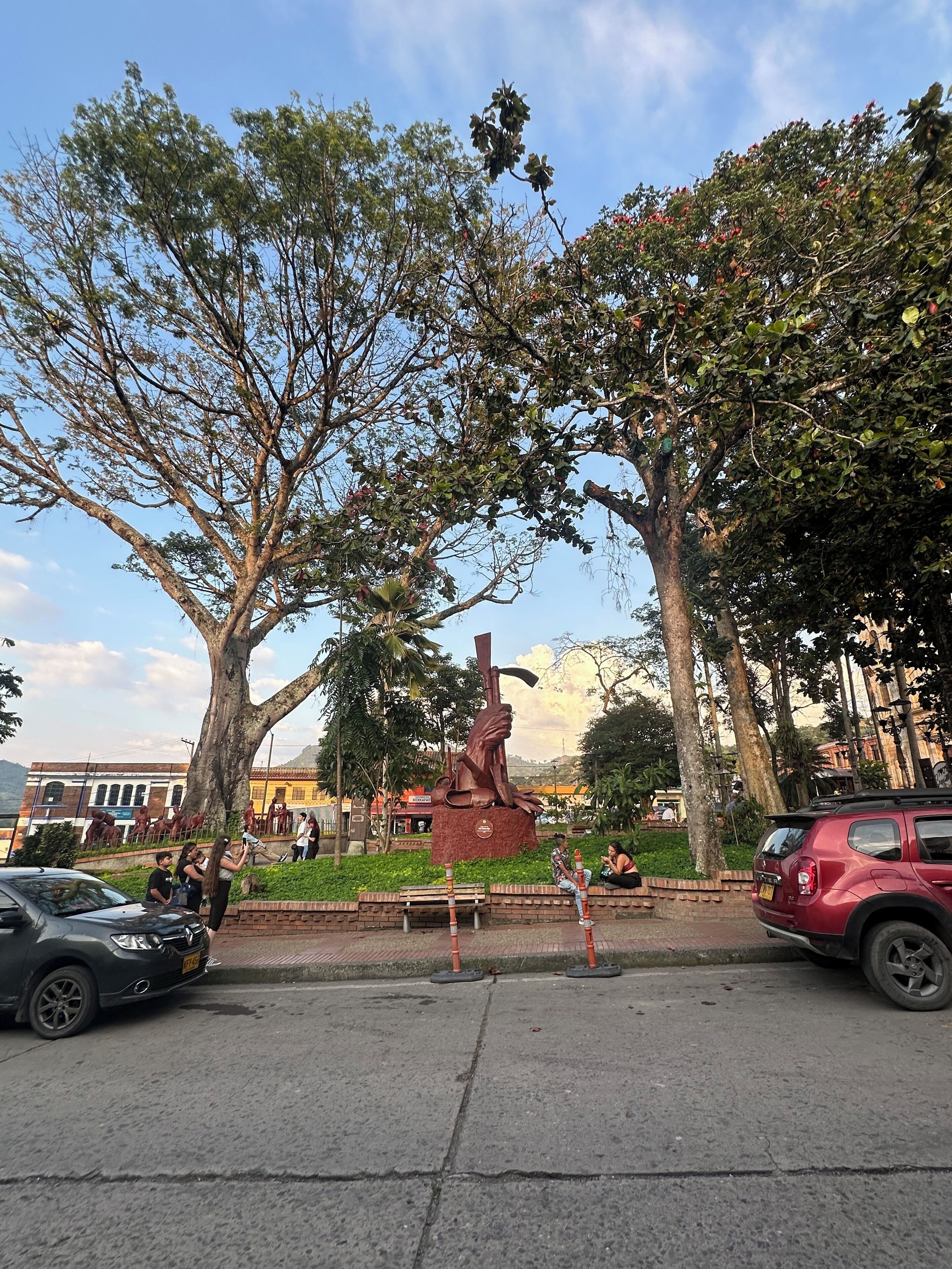 A park with a large tree, a statue, and a red car parked on the side of the road.