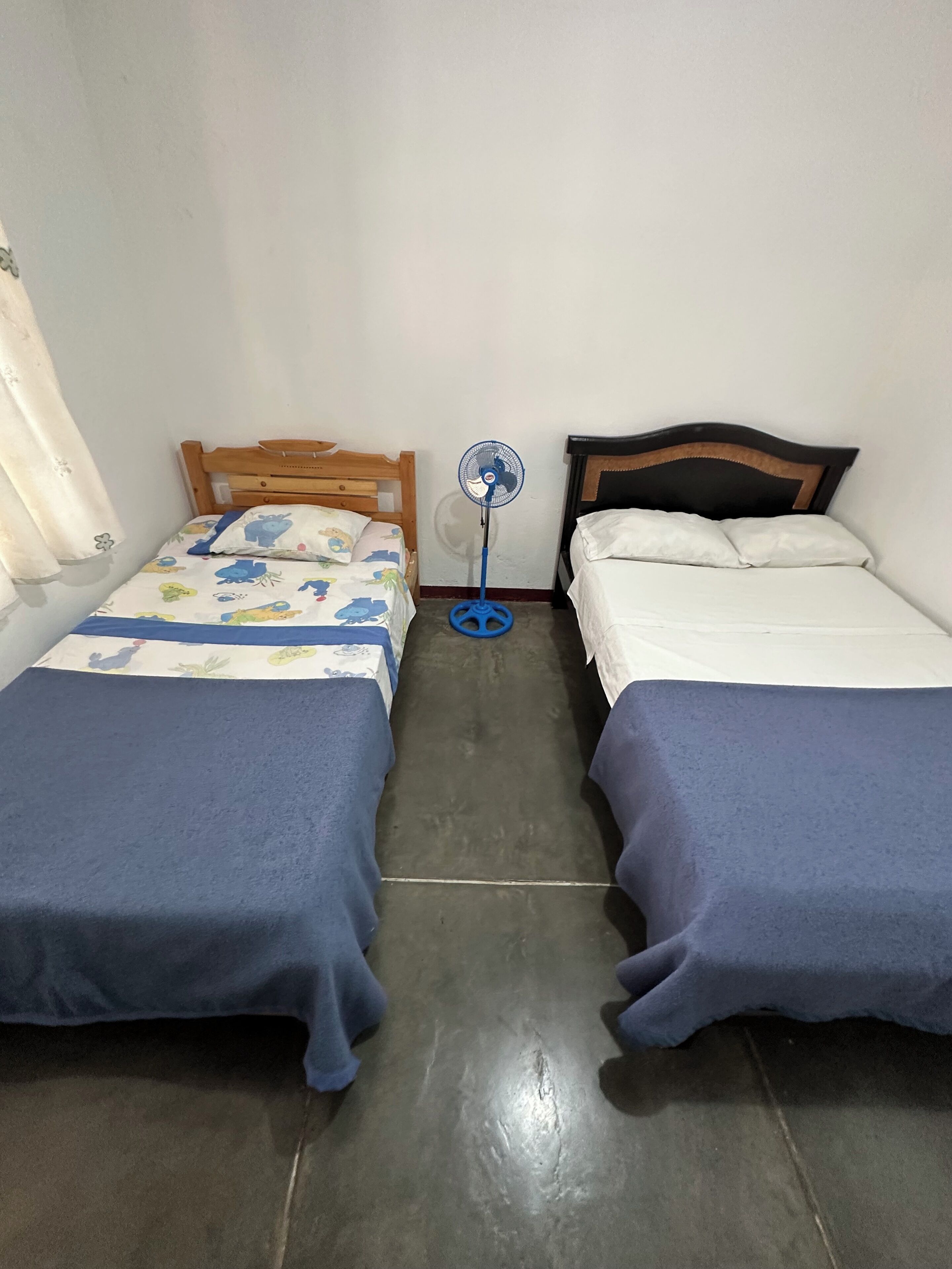 A room with two beds, each with a wooden headboard, a blue bedspread, and white pillows. A standing fan is between the beds.