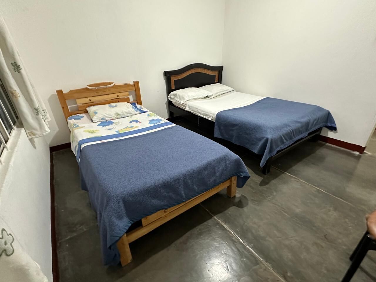 A room with two single beds, each with a wooden frame and blue bedding. The room has a tiled floor and white walls.