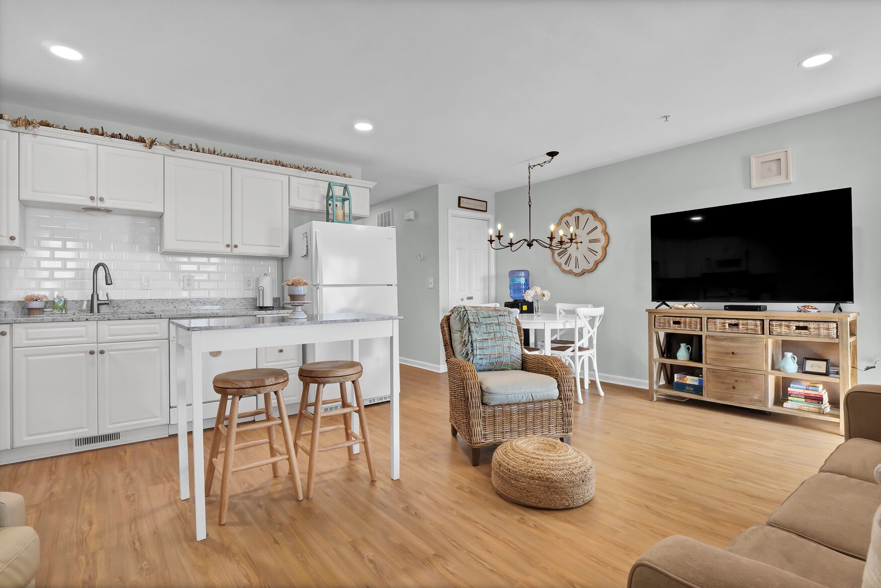 Une cuisine moderne avec des meubles blancs, un coin repas doté d’une chaise en osier et un salon avec une télévision et un meuble de rangement en bois.