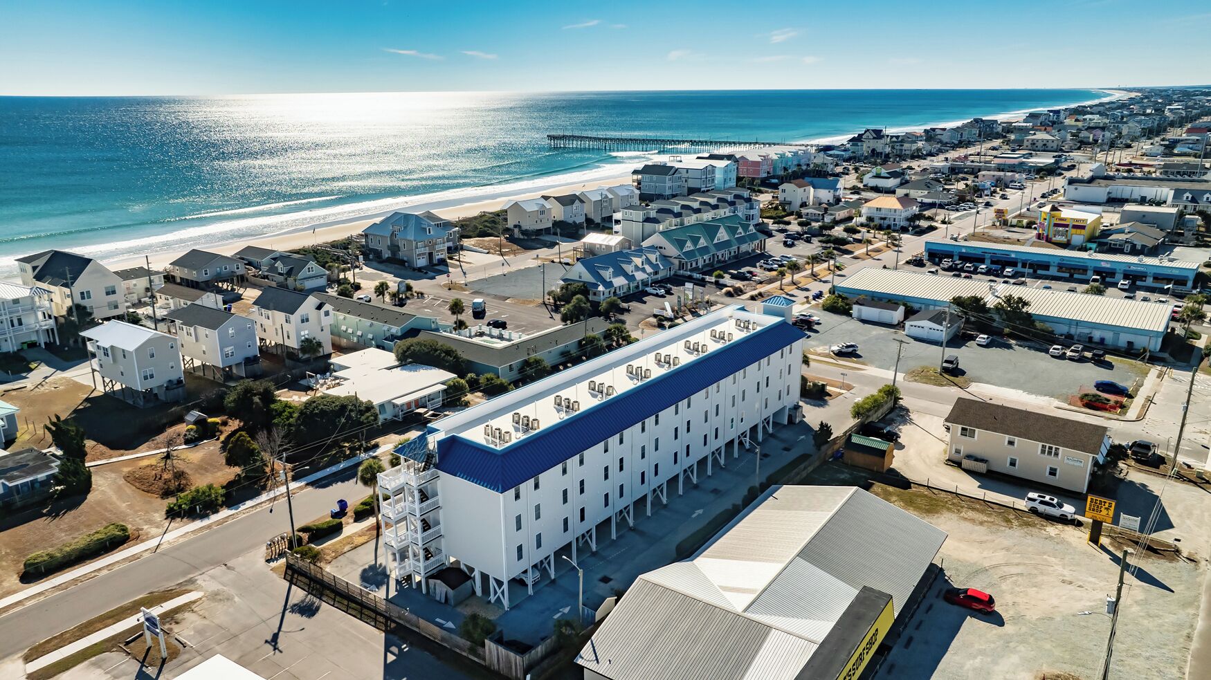 An aerial view of a coastal town with a prominent white building with a blue roof, surrounded by residential and commercial buildings, near a beach and ocean.