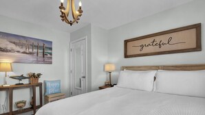 A bedroom with a large bed, a chandelier, a painting of birds on a fence, a 'grateful' sign, and a small bench with a 'happy' sign. - Perfect Surf City Stay: Walk to Beach, Dining, & Fun (Surf City)