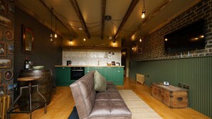 Un salon chaleureux avec un canapé en cuir, une table basse en bois et, au fond, une cuisine au style rustique.
