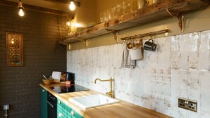 A kitchen with a wooden countertop, a sink, and a brick wall. There are hanging lights, a shelf with dishes, and a coffee maker on the counter.