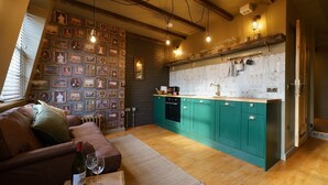 A cozy living room with a brown leather sofa, a wooden coffee table with a wine bottle and glasses, and a wall adorned with framed pictures. A kitchen with teal cabinets and a white backsplash is visible in the background.