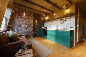 Un salon chaleureux avec un canapé en cuir marron, une table basse en bois sur laquelle se trouvent une bouteille de vin et des verres, et un mur décoré de tableaux encadrés. On aperçoit, au fond, une cuisine avec des meubles de couleur turquoise et un crédence blanche.