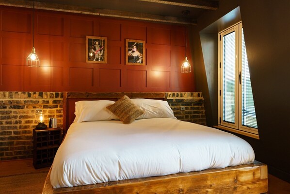 A bedroom with a wooden headboard, a large bed with white linens, two wall-mounted pendant lights, and a window with blinds. - GuestReady - Stylish Studio in Central London (London)