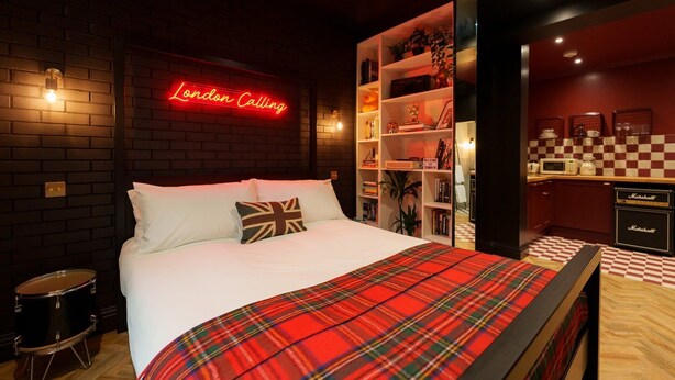 A hotel room with a bed, a neon sign reading "London Calling," a bookshelf, a drum set, and a checkered floor.