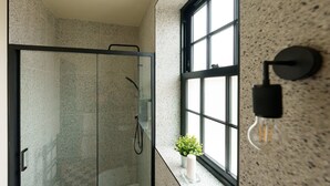 A modern bathroom with a glass shower enclosure, a window with a plant on the windowsill, and a wall-mounted light fixture.