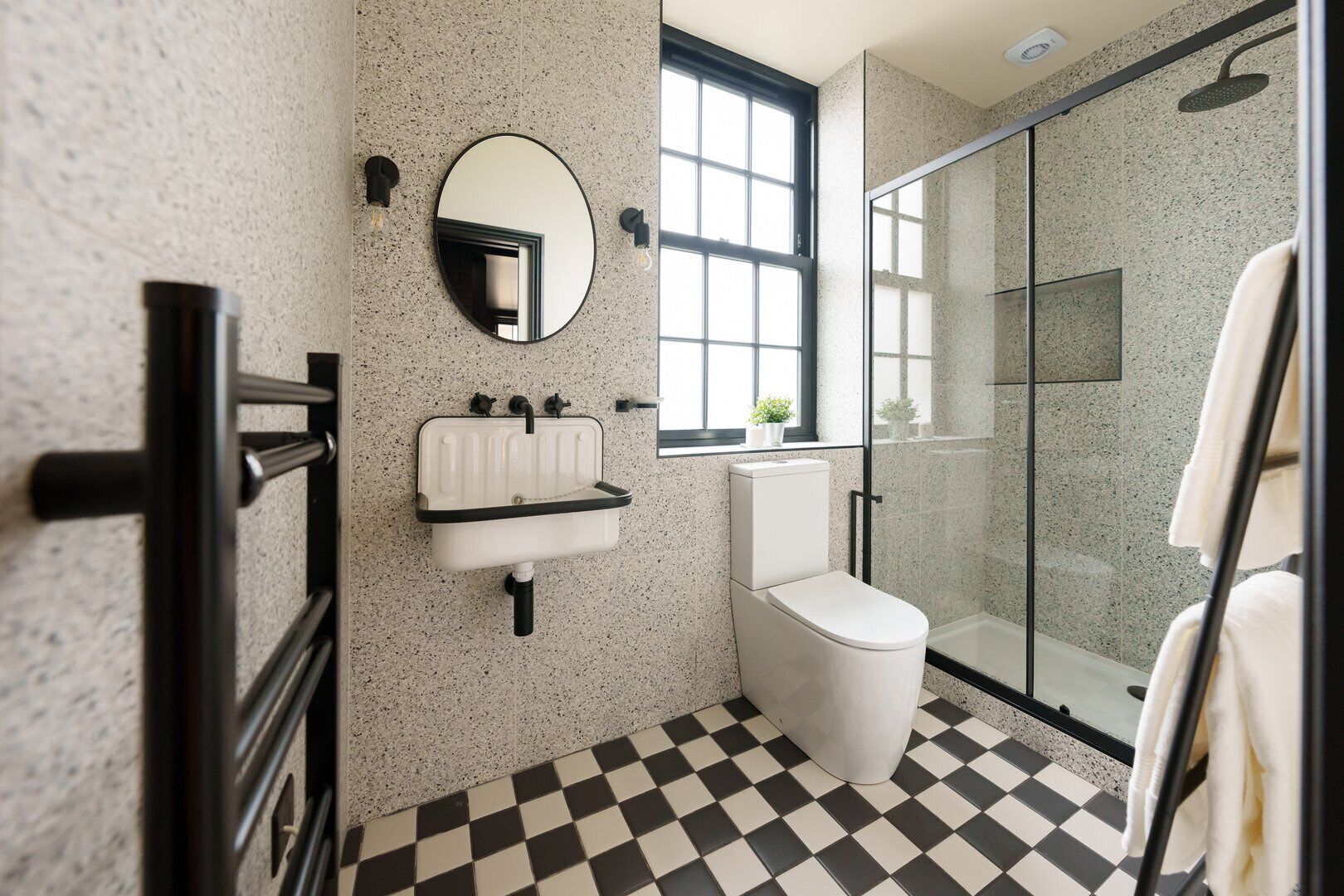 Ein Badezimmer mit Schachbrettmuster-Boden, einem weißen Waschbecken, einer Toilette, einer Duschkabine aus Glas und einem großen Fenster.