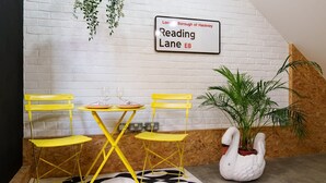 Ett rum med en vit tegelvägg, en skylt med texten ”Reading Lane E8”, ett gult bord med två stolar, en vit blomkruka formad som en svan och en svartvit mönstrad matta.
