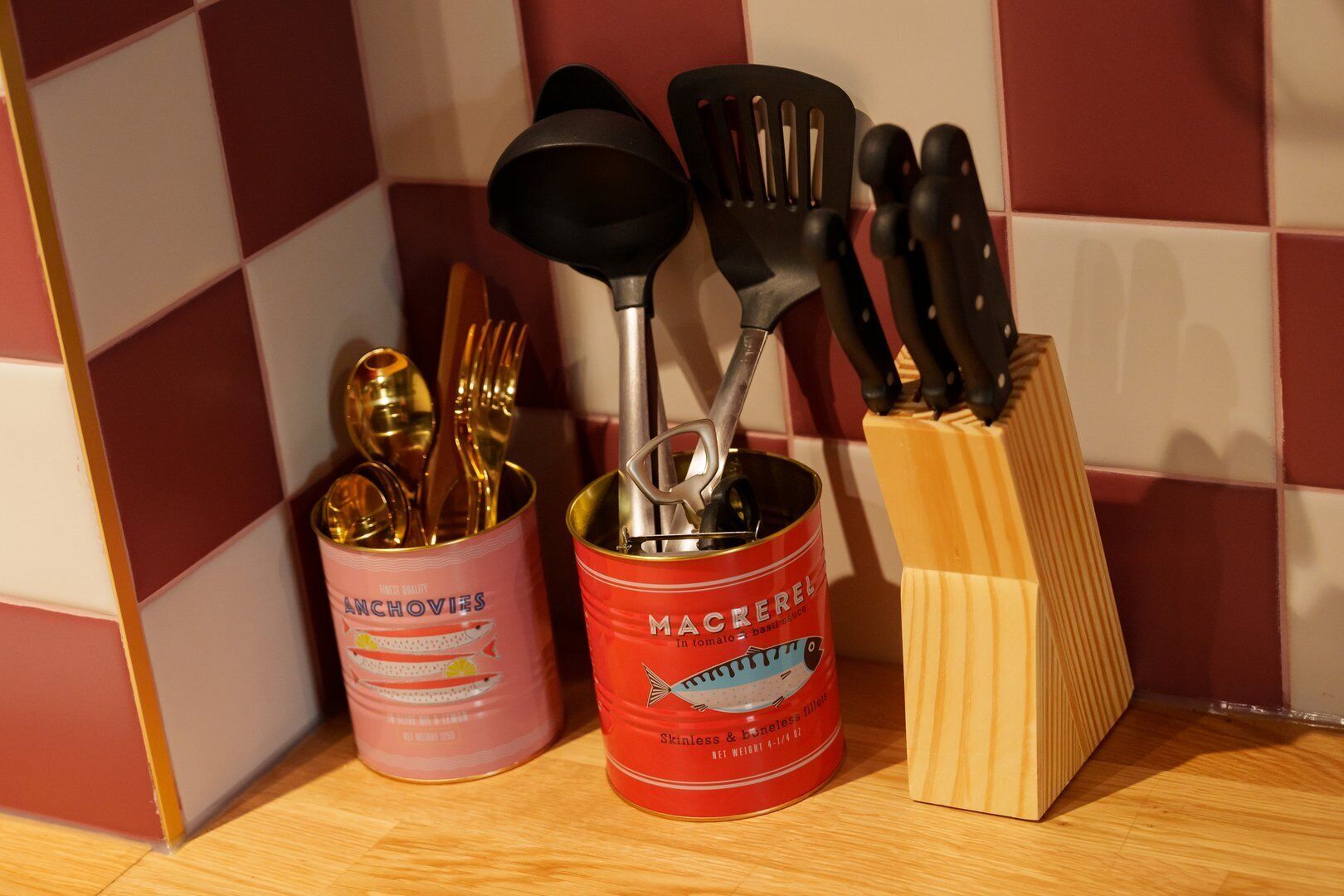 A kitchen shelf with a pink canister labeled 'ANCHOVIES', a red canister labeled 'MACKEREL', and a knife block.
